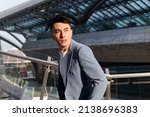 Small photo of Portrait of handsome Chinese young man in light blue suit and white undershirt walking and looking away with modern city buildings background in sunny day, side view of confident businessman.
