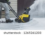A man in a black jacket and a gray pants is brushing white snow with the yellow electric snow thrower in winter