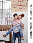 Small photo of Mischievous young woman riding piggy back on her husbands back as she brandishes a paint brush full of white paint while renovating the apartment