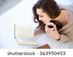 Cute lovely young woman reading book and drinking coffee on bed
