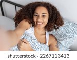 Cheerful young adult latin american woman taking selfie with phone while resting on bed in the morning. Happy millennial female sharing self portrait on social media network.