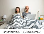 Unhappy or dissatisfied young couple in bed. Man and woman sit in modern bedroom after quarrel or dispute, girl depressed or sad, male with laptop computer