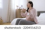 Small photo of unhappy asian female suffering heatstroke during pregnancy is fanning herself and loosening her hair while sitting with an arm akimbo in the bedroom.