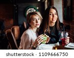 Two young women testing a cute fashion newly opened luxury restaurant, snacking by one green sandwich, looking at the camera
