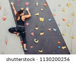 Female fitness professional climber training at bouldering gym. Muscular woman with athletic body dressed in black, climbing on artificial colourful rock wall. Active lifestyle and bouldering concept.