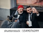 Small photo of Genial rich man in costume making selfie with caucasian beggar sitting on street, happy