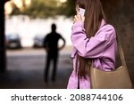 Criminal stalking woman, commiting crime while victim was walking alone, talking on phone in dark street. Caucasian young woman is looking back, afraid of male stranger person in the background