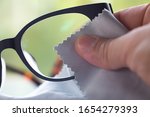 Small photo of Woman's right hand cleaning grey shortsighted or nearsighted eyeglasses by grey microfibre cleaning cloths, Bokeh green background, Close up & Macro shot, Selective focus, Optical concept