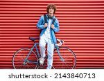 Stylish young male in casual clothes keeping hands on backpack straps while leaning on red metal wall and bike and looking at camera