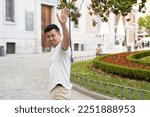Asian man looking sad while waving hand to say goodbye to someone outdoors in the street.