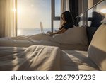 Small photo of Happy fresh relaxed asian woman resting sitting on bed in hotel room with two beds using laptop enjoying good morning after wakeup in cozy room with soft light from window.