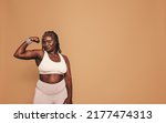 Small photo of Woman with dreadlocks looking at the camera while flexing her bicep. Mature woman standing against a brown background in sports clothing. Sporty black woman maintaining a fit lifestyle.