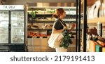 Small photo of Female shopper choosing food products from a shelf while carrying a bag with vegetables in a grocery store. Young woman doing some grocery shopping in a trendy supermarket.