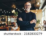 Small photo of Mature businessman standing in a co-working space. Happy mature businessman smiling at the camera while standing with his arms crossed. Experienced entrepreneur working in a modern workspace.