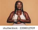 Small photo of Mature woman meditating with her eyes closed and her hands in prayer position. Black woman with dreadlocks practicing yoga in a studio. Happy middle-aged woman maintaining a healthy lifestyle.