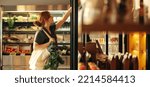 Small photo of Female customer choosing food products from a shelf while carrying a bag with vegetables in a grocery store. Young woman doing some grocery shopping in a trendy supermarket.