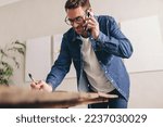Creative businessman writing notes during a phone call in his office. Happy young businessman making plans with his clients over the phone. Young businessman with glasses working remotely.