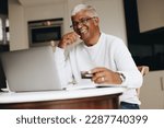 Small photo of Cheerful senior man smiling at the camera while shopping online at home. Happy mature man using his credit card to place an online order on a laptop. Senior man spoiling himself after retirement.