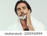 Young man appliying shaving cream to his beard, preparing himself for a smooth and comfortable shave. Man practicing his grooming routine, taking care of his facial hair to maintain a smooth look.