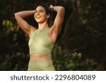 Small photo of Woman warming up for a morning workout outdoors. Happy sports woman tying her hair and preparing for yoga. Fit woman standing against a nature background.