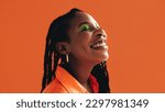 Small photo of African woman with makeup and face piercings smiling with her eyes closed. Black woman standing against an orange background.