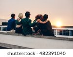 Rear view of young men and women relaxing together on rooftop and looking at sunset. Multiracial friends sitting on terrace in evening.