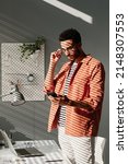 Small photo of Serious young mixed rac man in coral shirt adjusting eyeglasses and checking smartphone in office with minimalistic interior