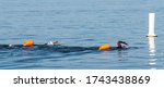 Two black female triatholon swimmers with orange flotation devices for safety are about to pass a buoy while training in the sea.