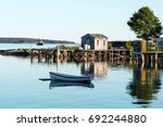 Small photo of View of Bass Harbor Maine from the deck of Thurston's Lobster pound with a dock, lobster traps, boat house, tree and row boat reflections visible. A fishing boat is in the background