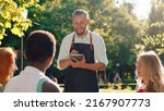 Small photo of A very good looking waiter with his hair slicked back, is taking down orders in his notebook, he s talking to three women taking their orders and discussing while being in a park scenery