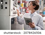 Small photo of Portrait of female factory worker wearing glasses and white robe, pressing machine buttons, using tablet for quality control and logistic purposes at polymer plastic manufacturing