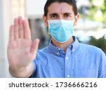 STOP COVID-19 Man wearing surgical mask on face doing STOP gesture looking at camera outdoors. Young man with face mask showing open hand palm at the camera against Coronavirus disease 2019.