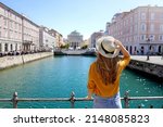 Travel in Trieste, Italy. Back view of pretty girl holding hat looking at Sant Antonio Taumaturgo church on Grand Canal in Trieste, Italy. Beautiful young woman visiting Europe.