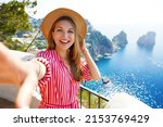 Small photo of Fashion beauty woman taking a self portrait smiling at the camera in Capri Island with Faraglioni sea stack and blue crystalline water on the background, Capri, Italy