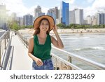 Small photo of Stylish girl with hat walking along promenade on windy day with skyline skyscrapers on the background, Santos, Brazil