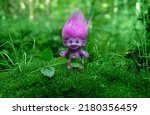 Small photo of cute toy troll close up on natural abstract green background. spring summer season. little purple troll with ruffled violet hair in mystery forest. fairytale, magic atmosphere
