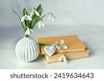Bouquet of Snowdrop flowers, heart decor and books on table, abstract light background. symbol of spring season. Relaxation, reading time, harmony of nature. romantic composition. template for design