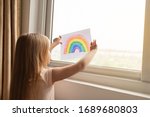 Kid painting rainbow during Covid-19 quarantine at home. Girl near window. Stay at home Social media campaign for coronavirus prevention, let's all be well, hope during coronavirus pandemic concept