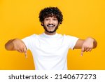 Amazed indian or arabian cheerful young man in white basic t-shirt looks at the camera and points fingers down at space for your presentation, stands on isolated orange color background, smiling