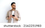 Small photo of Panoramic photo of a joyful handsome indian or arabian guy, holding smartphone in hand, chatting online, browsing internet, looking happily to the side, standing on isolated white background, smiling