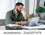 Small photo of Tired exhausted arabic or indian man, office worker, manager or freelancer, sitting at his desk, tired of working in a laptop, overworked, having a headache, closed his eyes, needs rest and break