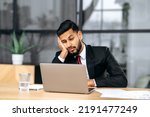 Small photo of Fatigue, chronic sleep deprivation. Sleepy arabian or indian business man in a suit, sit at workplace in office with a laptop, put his head on his hand, fell asleep during work, feels tired