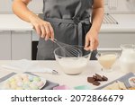 Small photo of Women's hands whisk cream, cream, mousse, dessert in a transparent bowl against the background of a white kitchen. Grey apron. Marshmallows, cream, milk, chocolate. Side view. Confectionery.