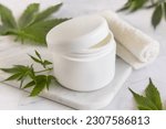 Opened white cream jar with a lid near green cannabis sativa leaves on a marble table. Cosmetic Mockup, Copy space. Organic skincare beauty product. Eco friendly body or hand cream with hemp