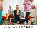 Stressed out mother sitting on floor in middle of toys while children naughty running around her at room. Woman alone burnout with kids. Family home with chaos, mess. Motion blur for speed, real life.