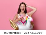 Happy excited teen girl with open mouth and inflatable unicorn ring hold phone in one hand and hold head with the other, young tik tok blogger, good news wear swimsuit isolated on pink background