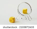Small photo of Yellow dice with sad expression looking in the mirror and seeing himself smiling - Concept of person who hides his pain behind his smile