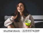 Brunette woman eat bucket of pistachio ice cream and watch tv, change channel with remote control sitting on bed in bedroom. Happy laughing girl watch comedy movie, have fun at home.