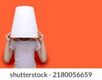 The child put a bucket on his head, sitting on an orange background. Funny child hid in a white bucket. The child hides from the parents. Free space for text