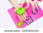 Small photo of The child plays and sculpts figures from soft green plasticine on a pink board. The child's hands hold plasticine. The child plays and has fun with plasticine. Creative development of the child